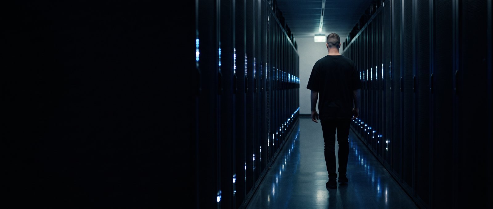 A man walks down a narrow corridor between rows of server cabinets lit by small blue lights.