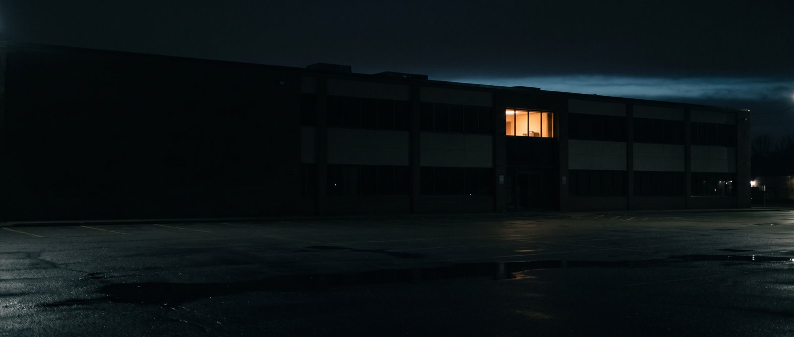 A dark two-storey office building at dusk with a single window lit on the upper floor, across an empty wet car park.