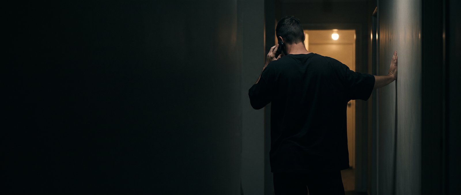 A man in a black t-shirt holds a phone to his ear, leaning on the wall of a dark hallway that opens onto a lit doorway.