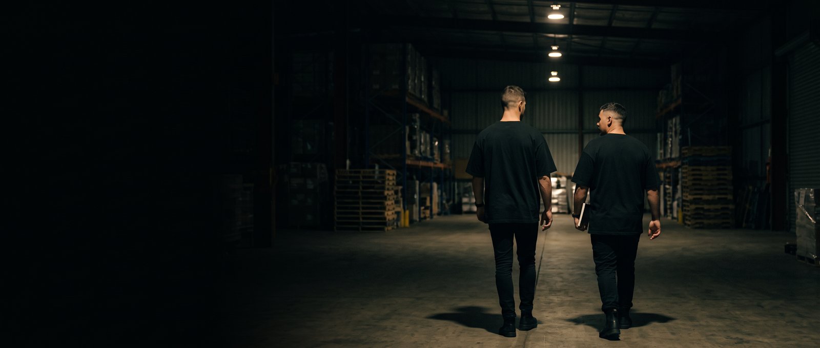 Two men in black t-shirts walk down the centre aisle of a dimly lit warehouse lined with pallet racking.